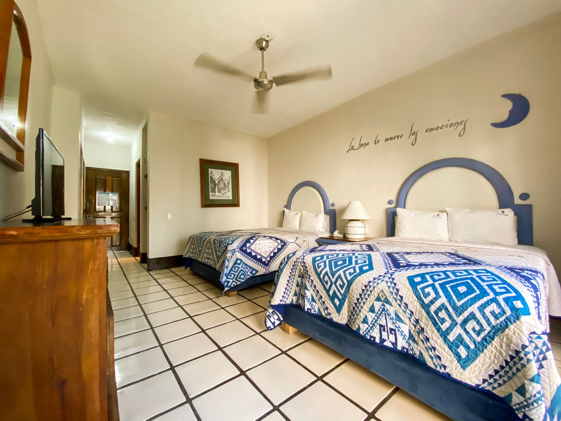 Bedroom with two beds, blue and white patterned quilts, tile floor, ceiling fan, TV, and wall art.