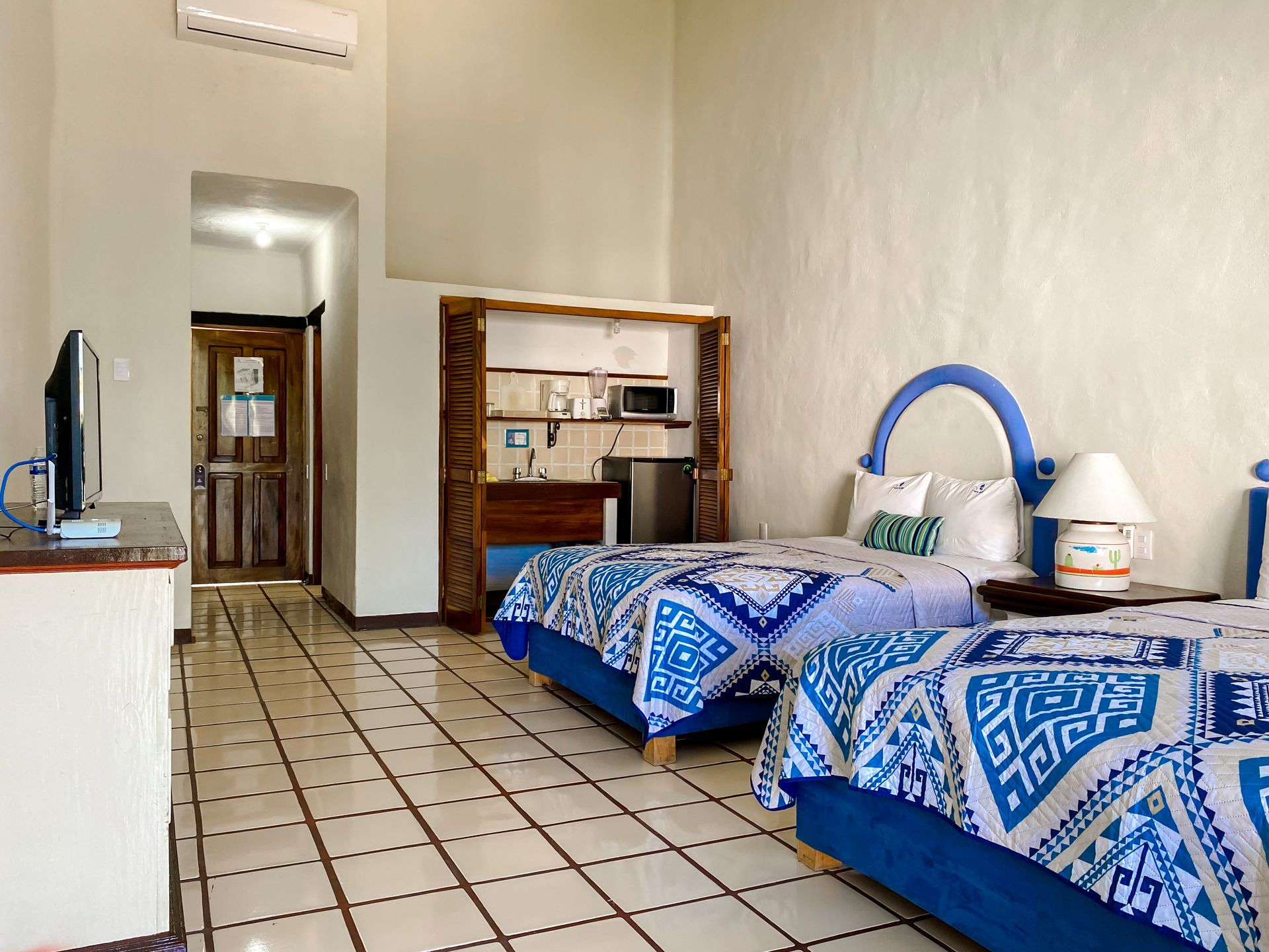 Hotel room with two beds, kitchenette, TV, and open doorway to another room.