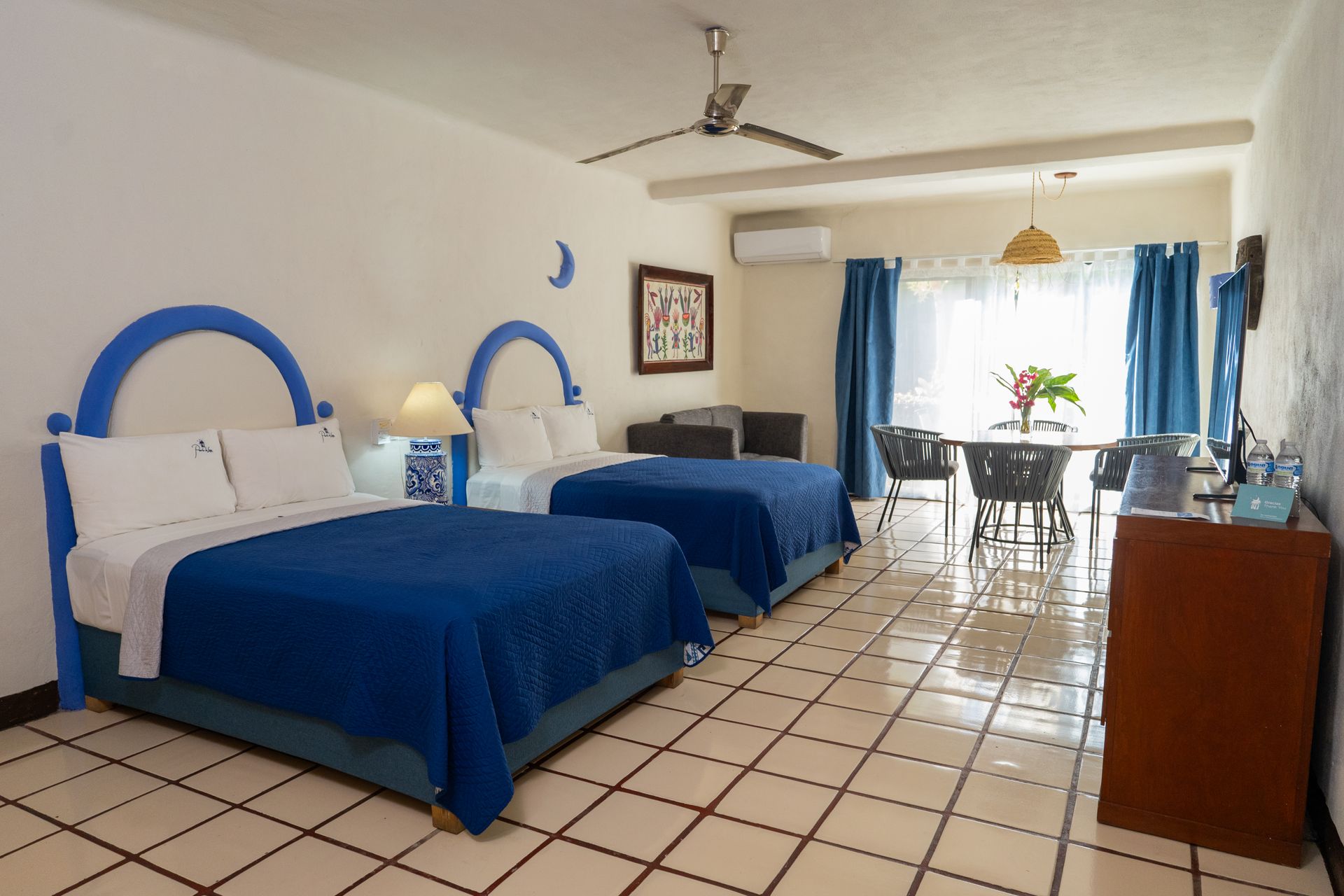 Hotel room with two twin beds, blue accents, dining area, and tan tile floor.