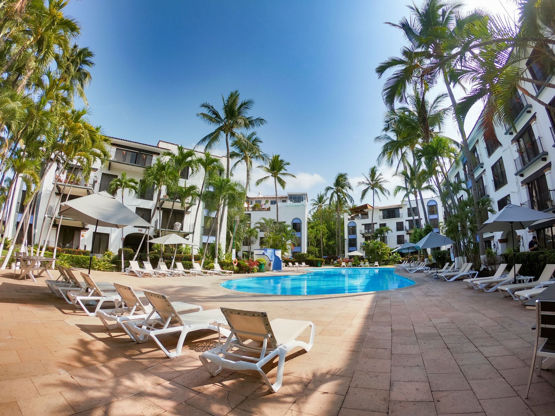 Swimming pool and lounge chairs at a tropical resort, with palm trees and white buildings under a blue sky.