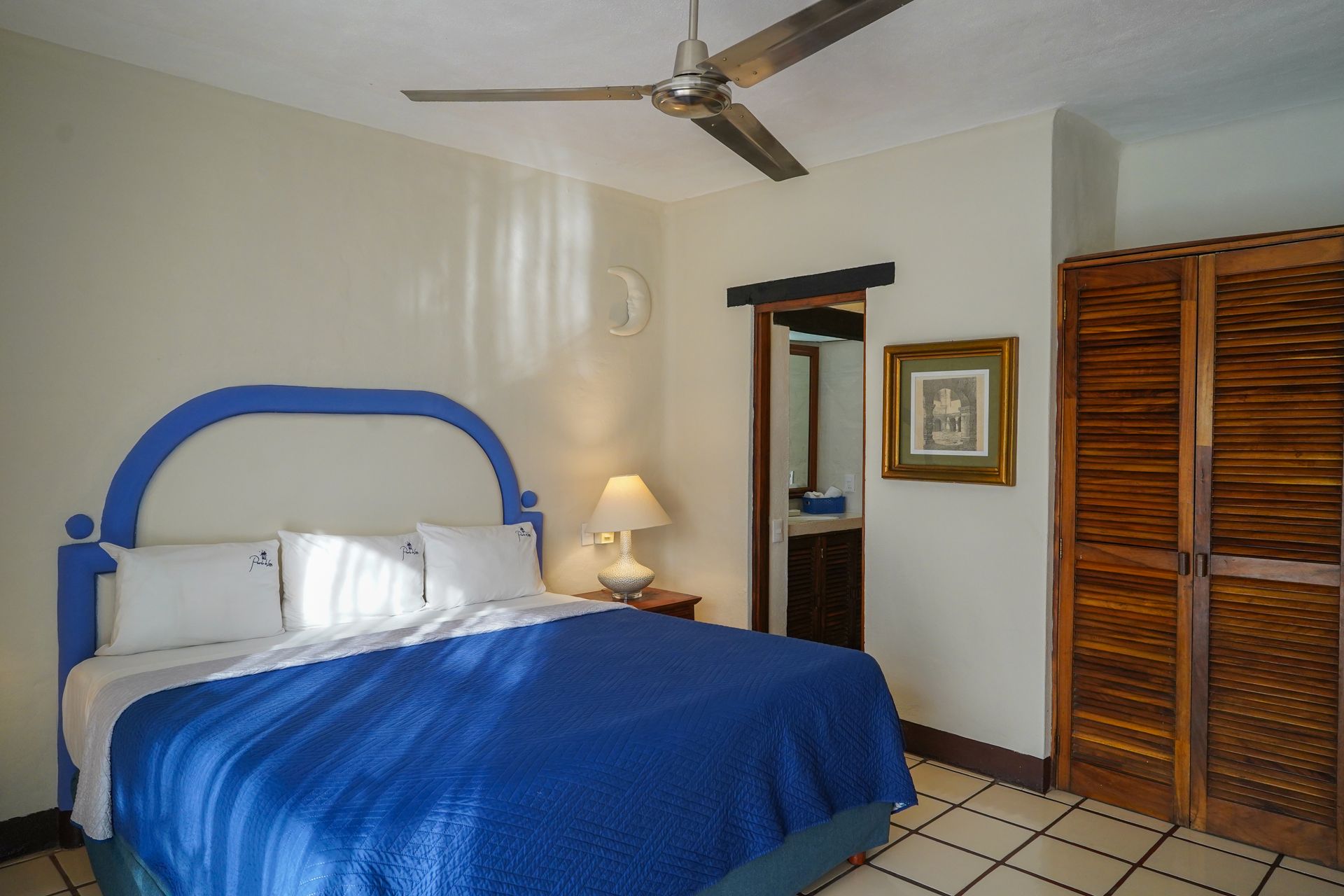 Bedroom with blue bedspread, headboard, and wooden closet; bathroom visible through doorway.