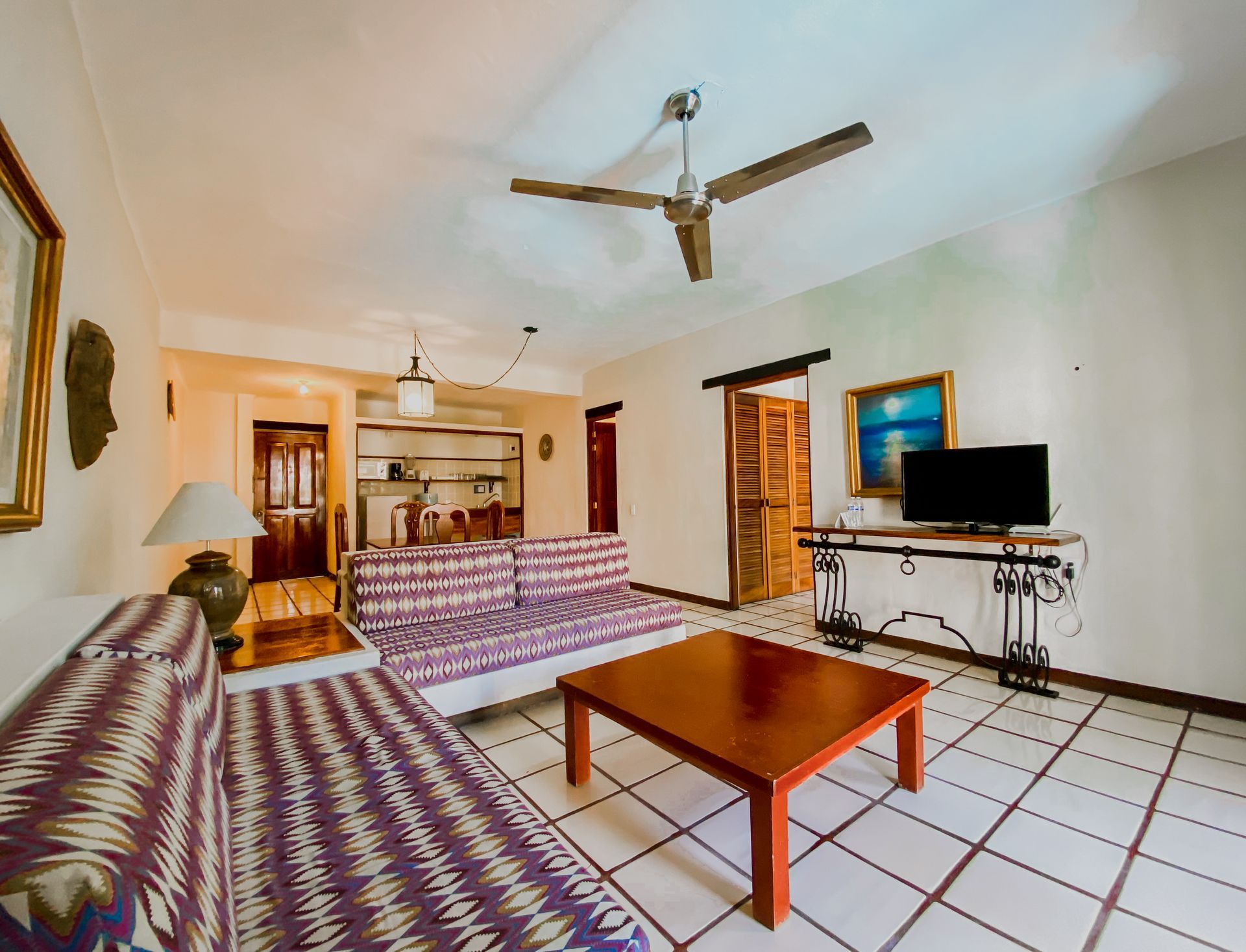 Living room with couches, coffee table, TV, and ceiling fan. White walls, tile floor, and wooden doors.