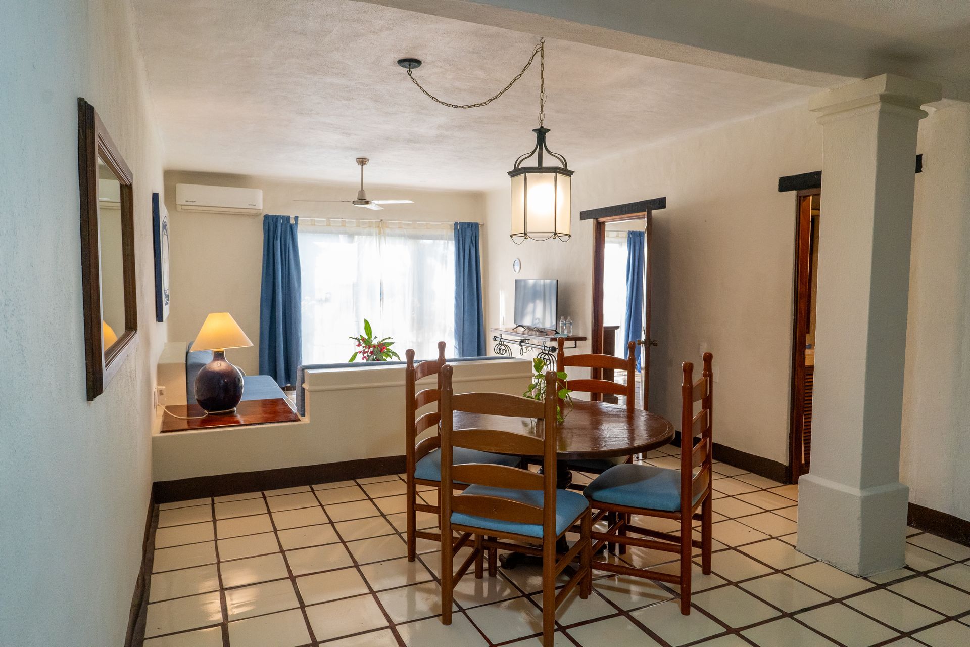 Dining area with round table, four chairs, and a hanging lantern light. Window with blue curtains.