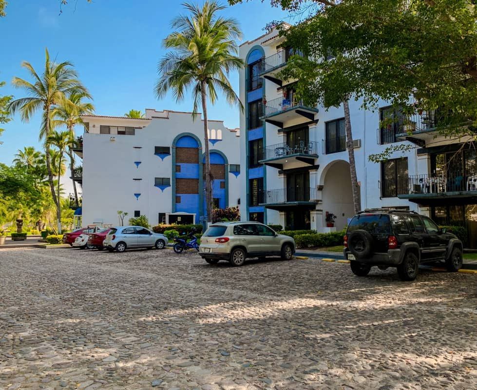 Cars parked in a cobblestone lot in front of a white and blue building with palm trees.