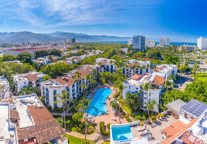 Aerial view of a resort with blue pools, white buildings, and lush green trees near the ocean.