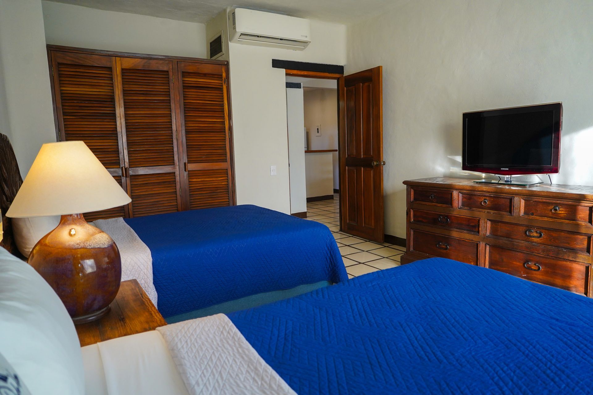 Bedroom with two twin beds, blue covers, wood dresser and closet, TV, and lamp.