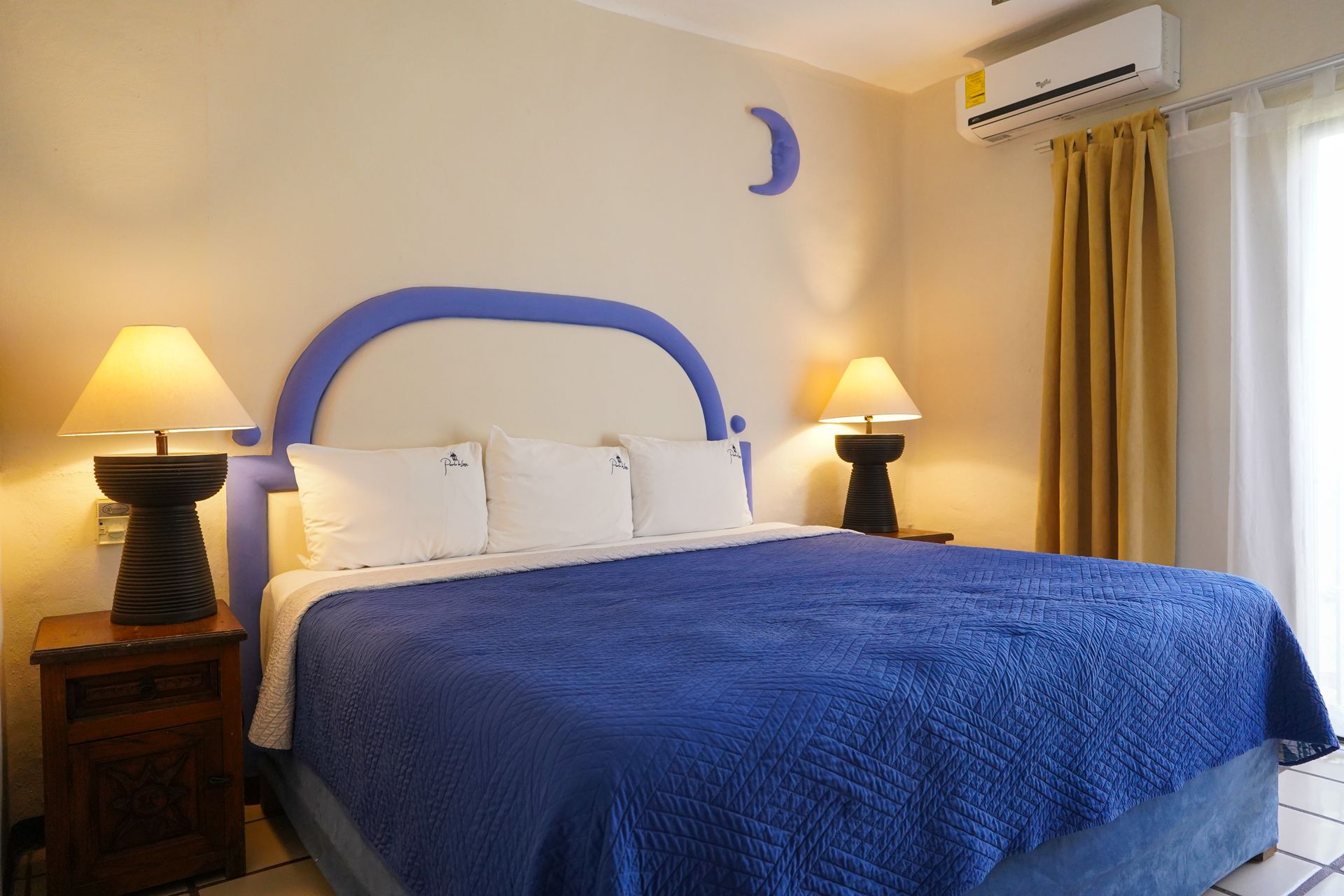 Bedroom with blue bedspread, headboard, lamps, and air conditioner.