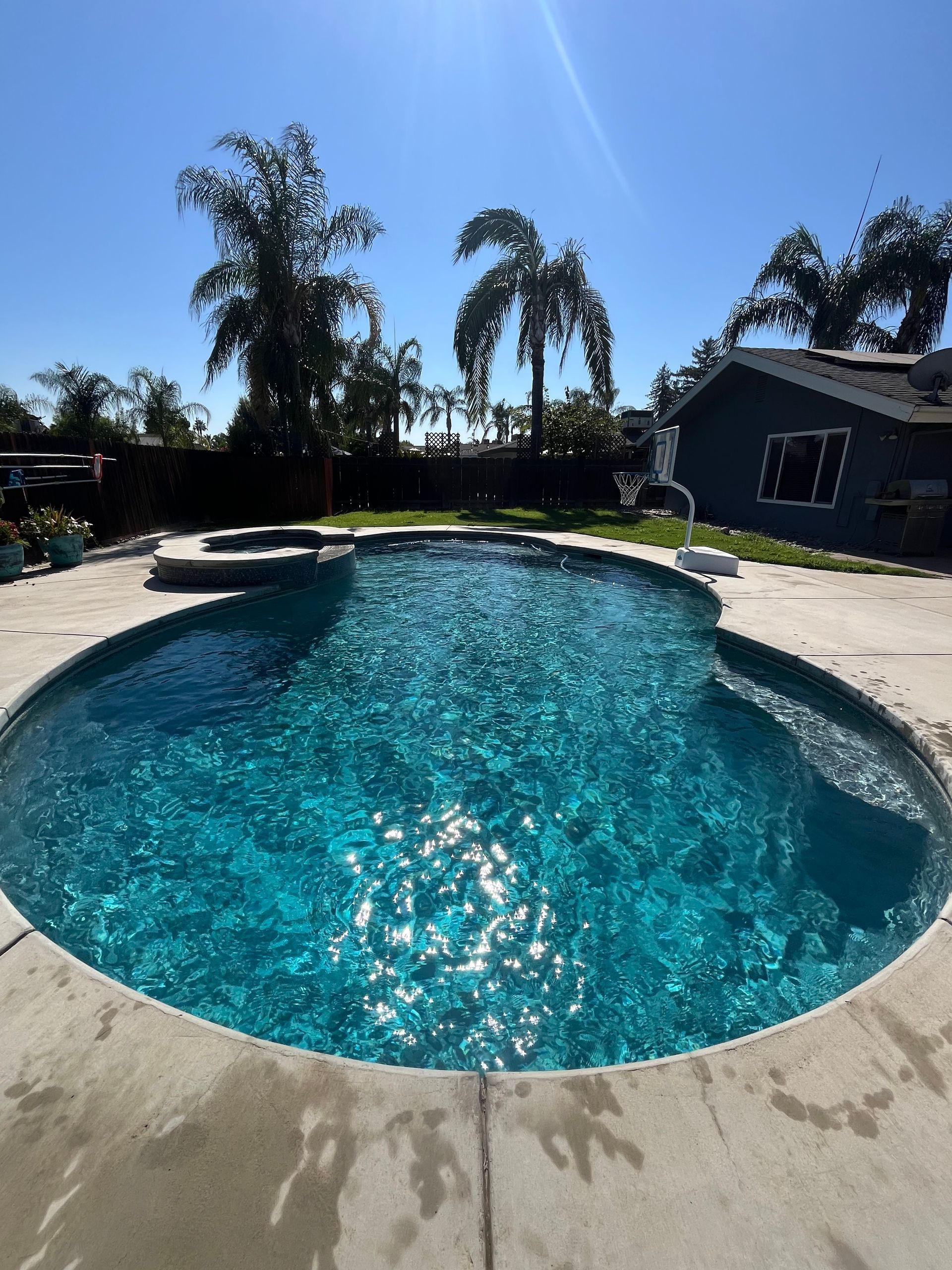A swimming pool with turquoise water, surrounded by concrete, palm trees, and a blue sky.