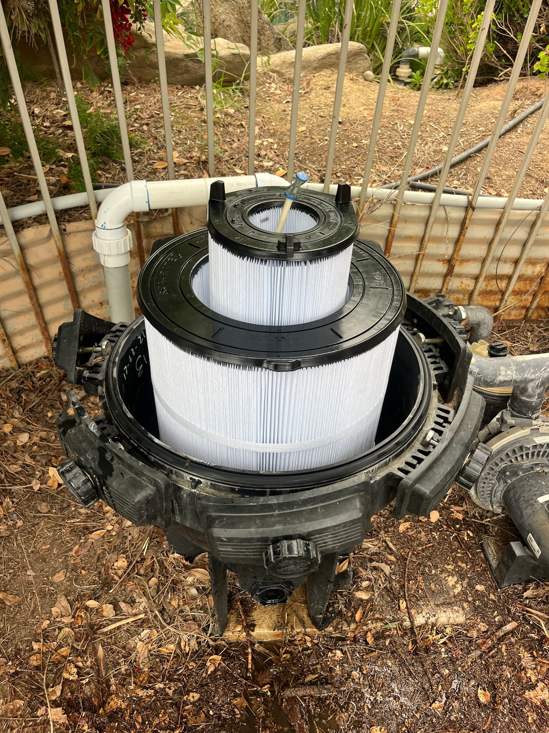 Open pool filter with cylindrical, pleated filter cartridge. Black housing, white filter media, outdoors.