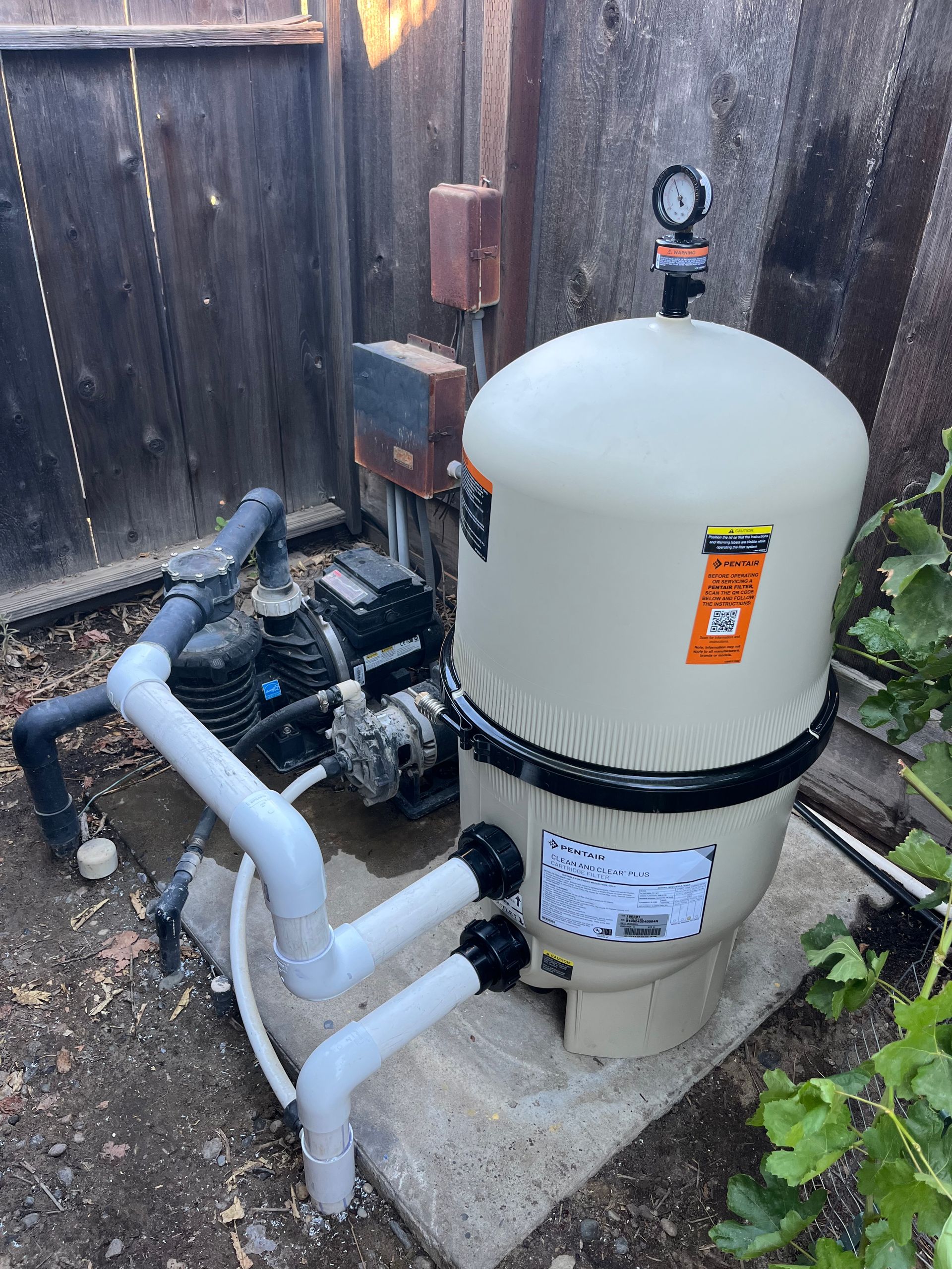 Pool filter system outdoors, tan filter tank, pump, black pipes, wooden fence background.
