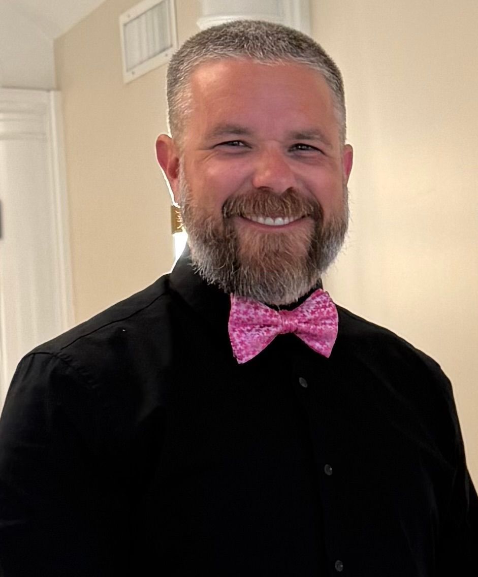 Man with graying beard, wearing a black shirt and pink bow tie, smiling.