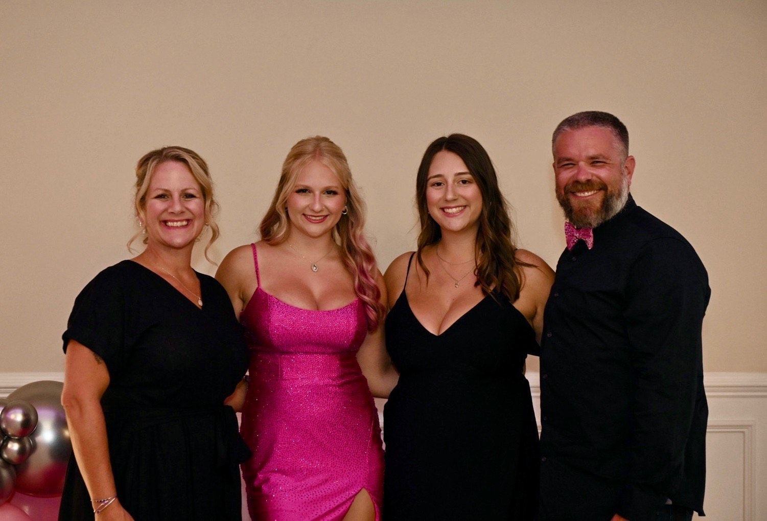 Family of four in formal attire posing for a photo. Two women in dresses, two adults smiling. Balloons in background.