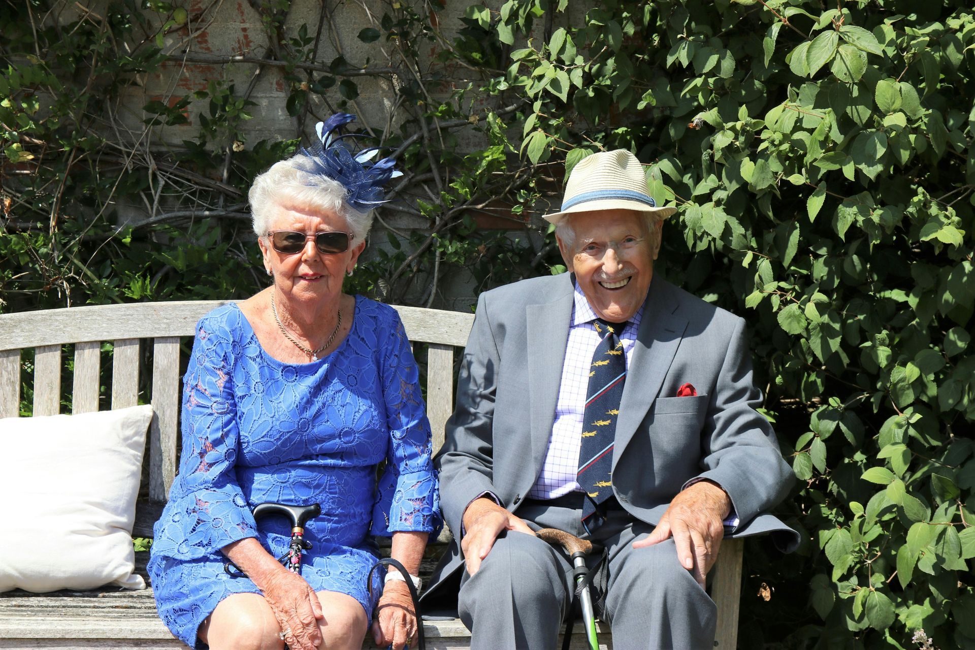 Elderly couple smiling, sitting on a bench outdoors. Woman in blue dress and hat, man in gray suit and fedora.