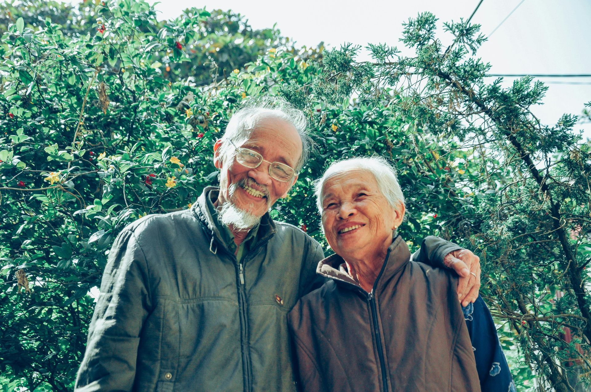 Elderly couple smiling, hugging, with green foliage in the background.