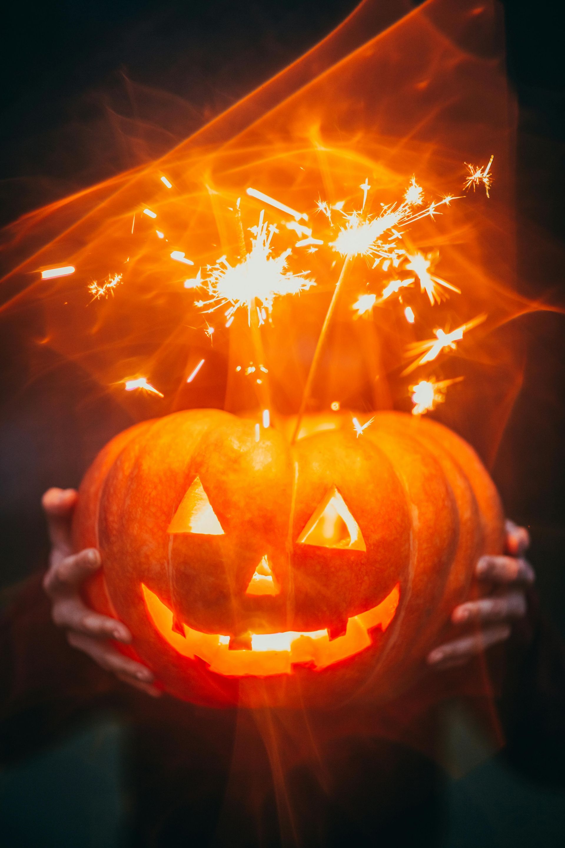 Carved jack-o'-lantern held by hands, lit with sparklers and a smiling face. Glowing orange light.