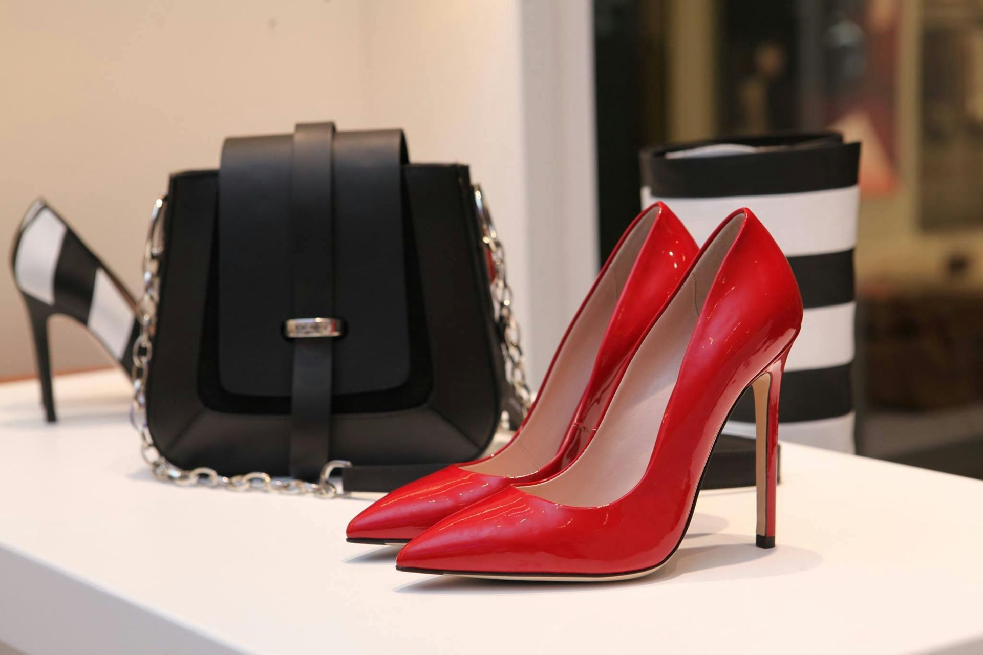 Red high heels, black handbag, and a striped cylinder on a white shelf.