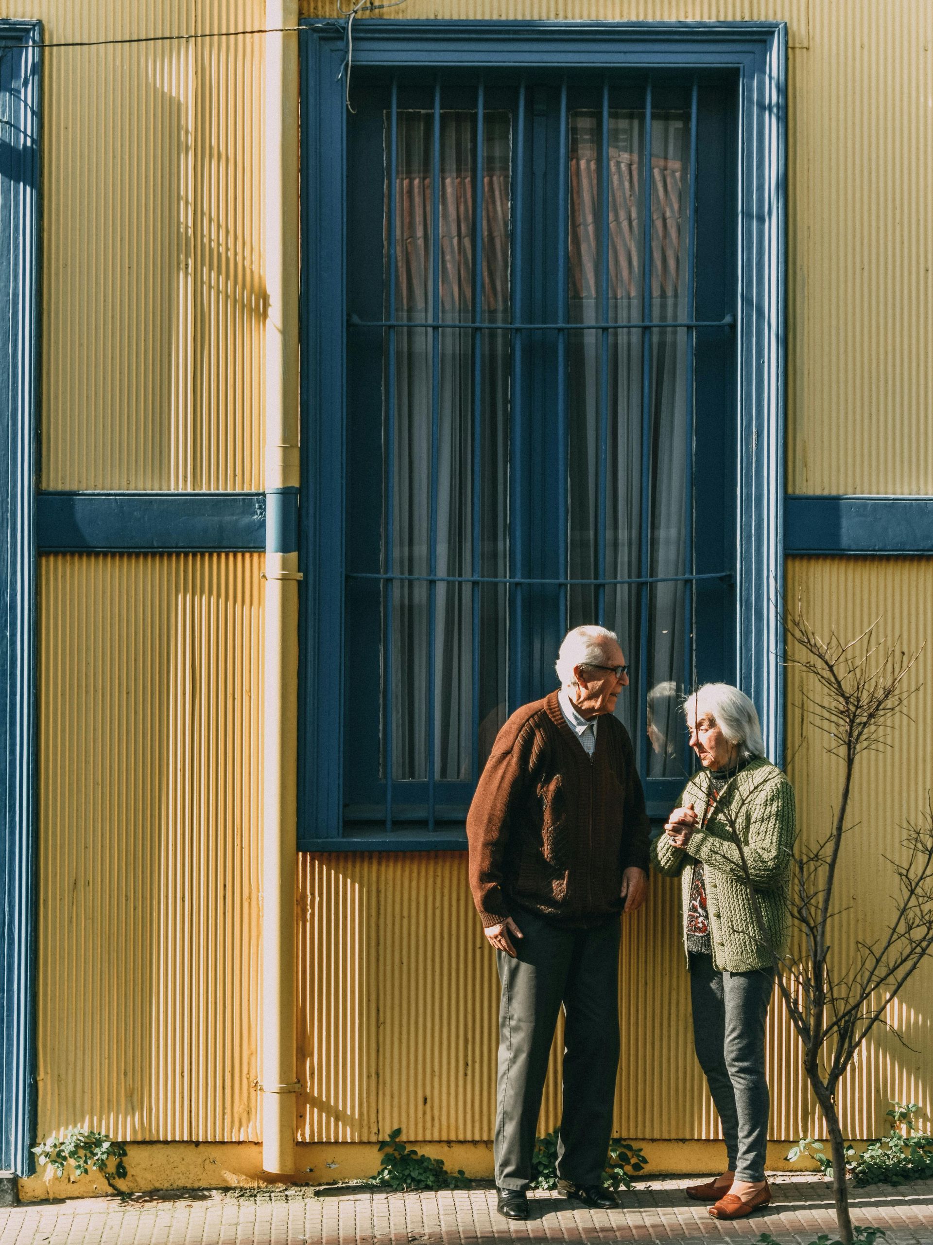 Elderly couple standing by a yellow and blue building, near a window with bars.