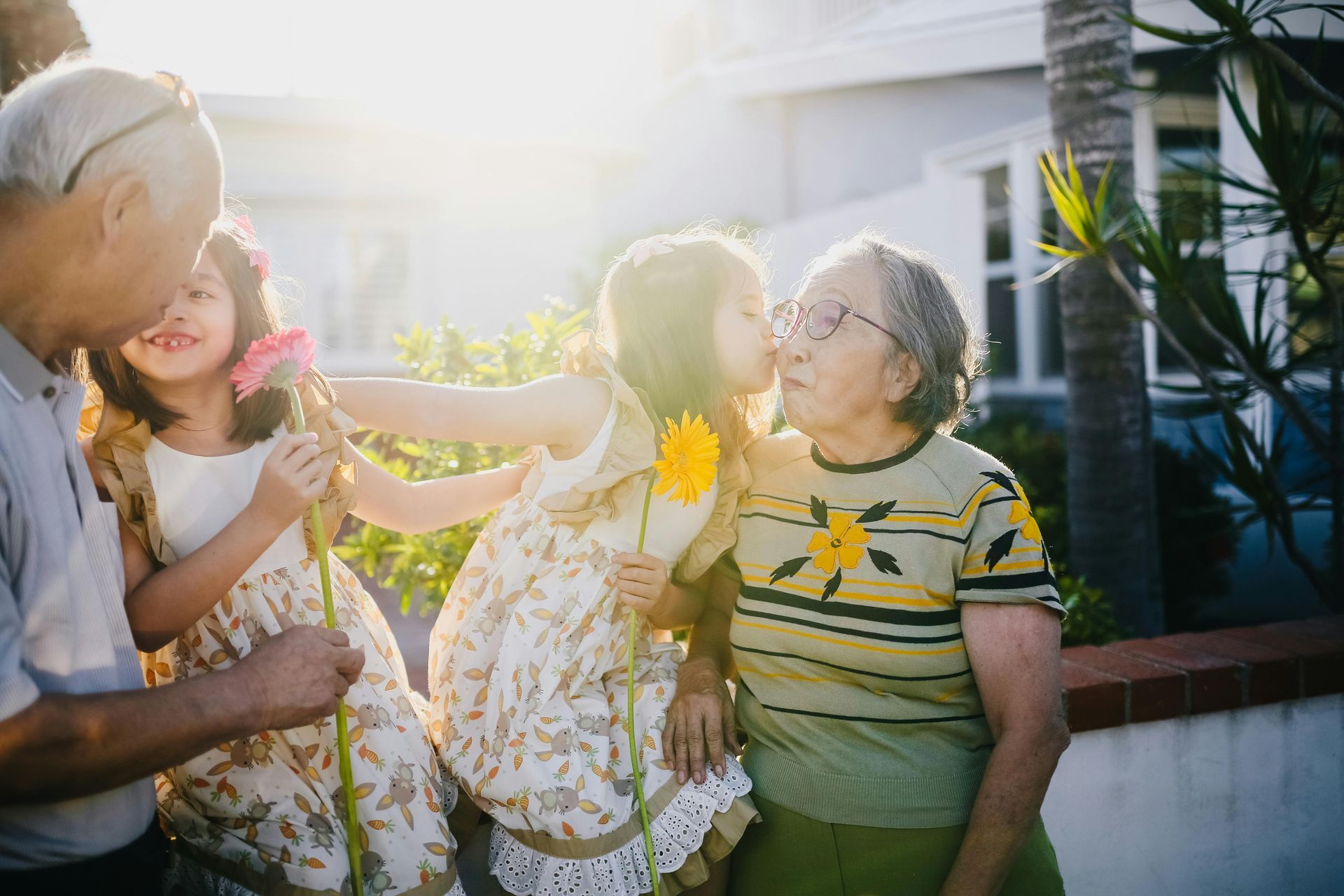 Grandparents with grandchildren, sunlit outdoors, give flowers and kisses.