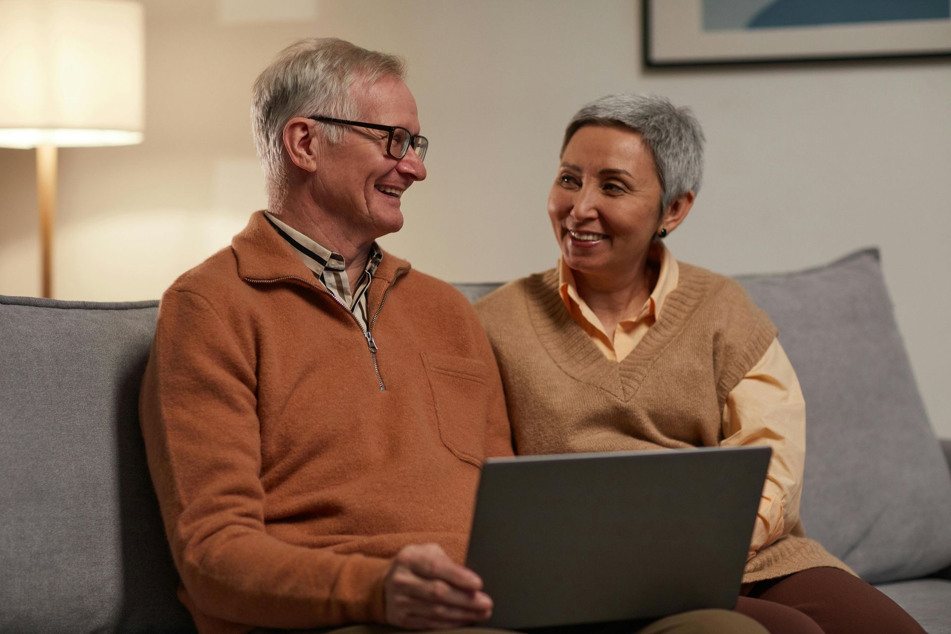Older couple sitting on a couch, looking at a laptop, smiling.