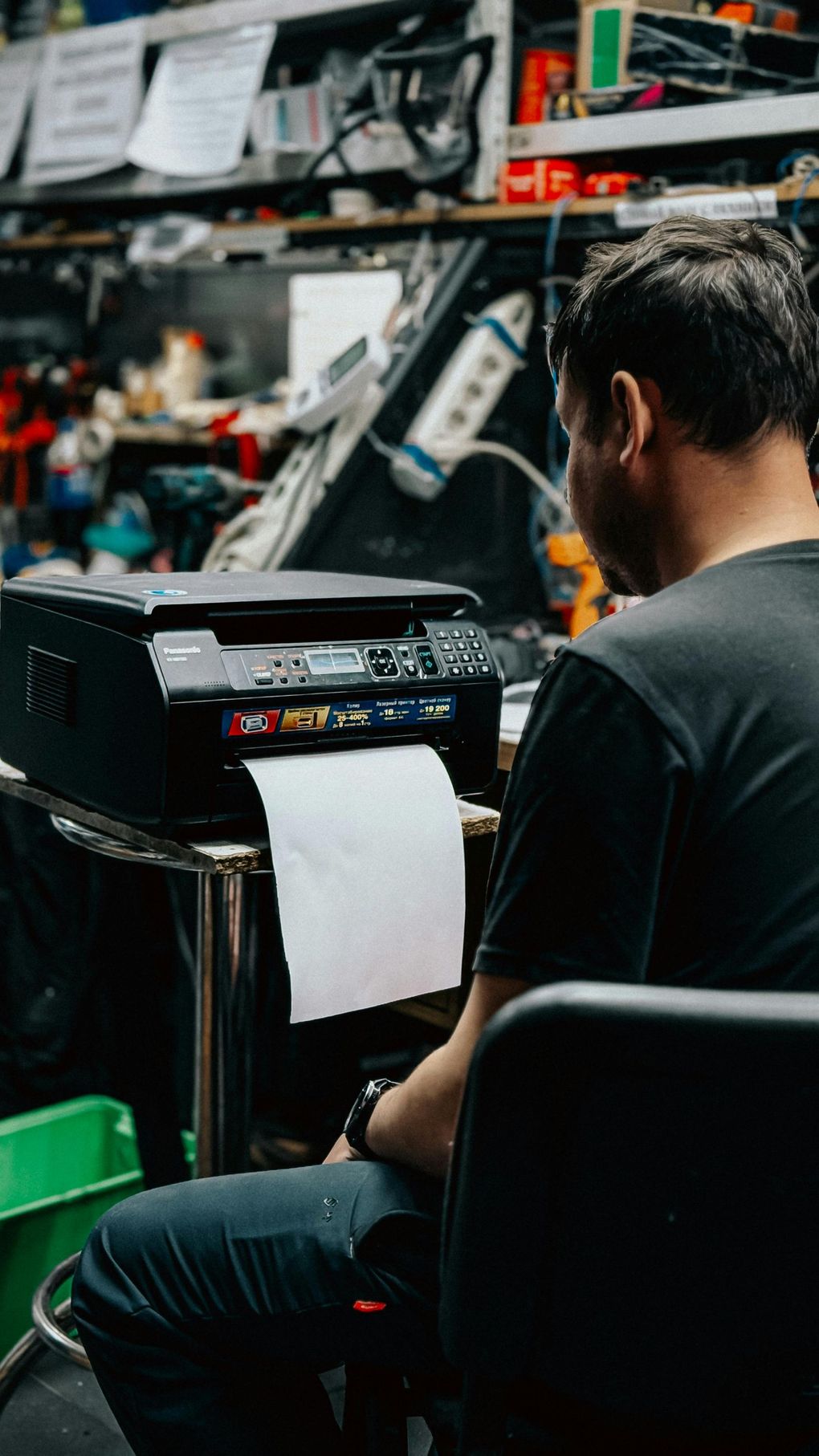 Man seated at a printer in a cluttered workshop, holding paper as it prints.