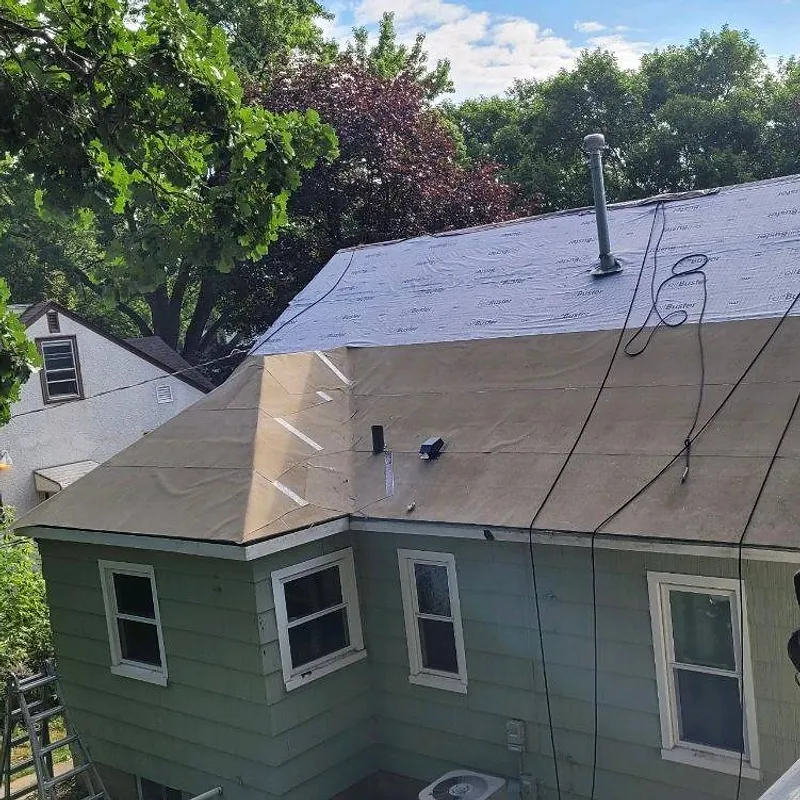 House with plywood and partial roofing; green siding, several windows. Roof work in progress.
