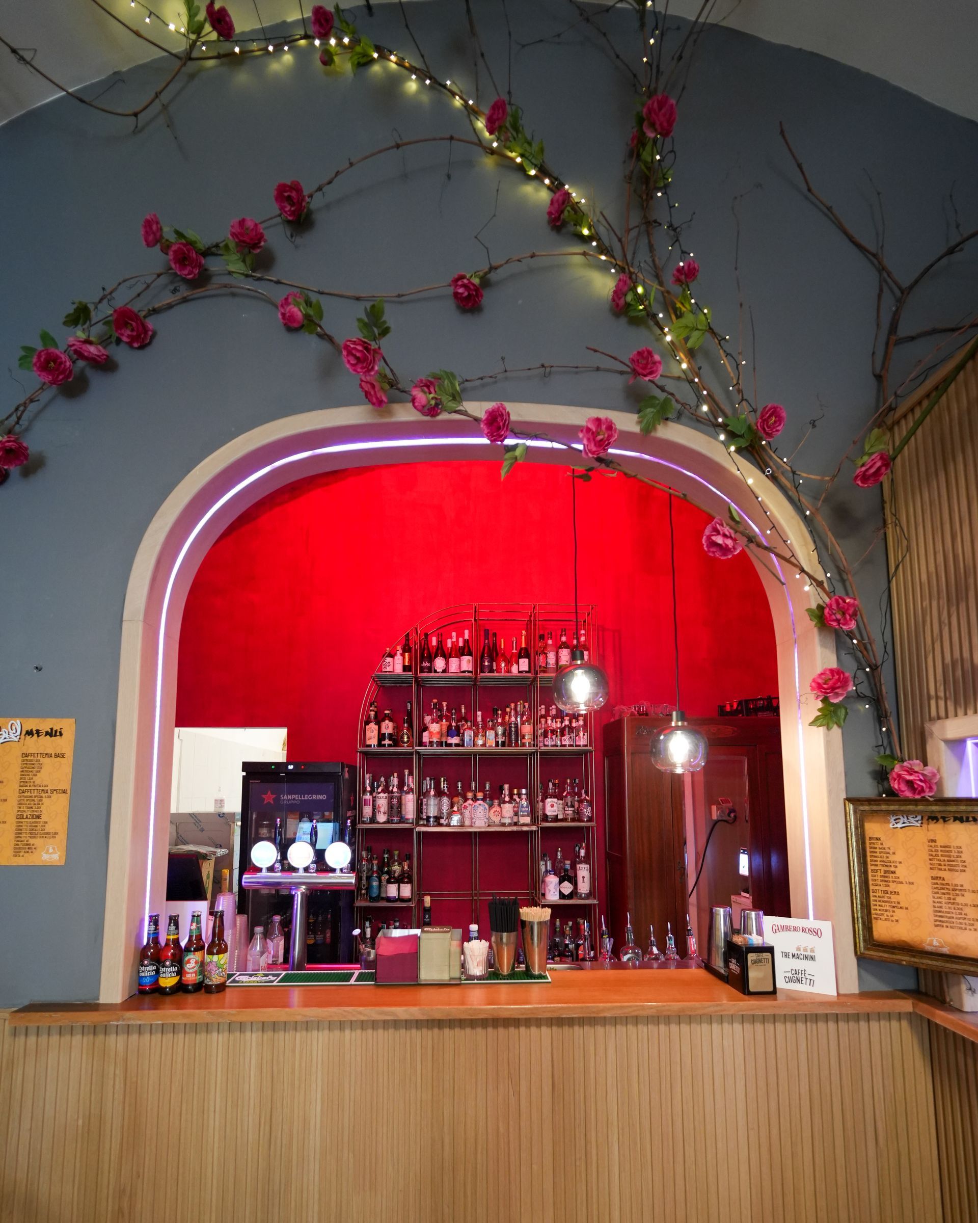 Bar con parete rossa sullo sfondo, bancone in legno, scaffali pieni di liquori e ghirlanda di rose.