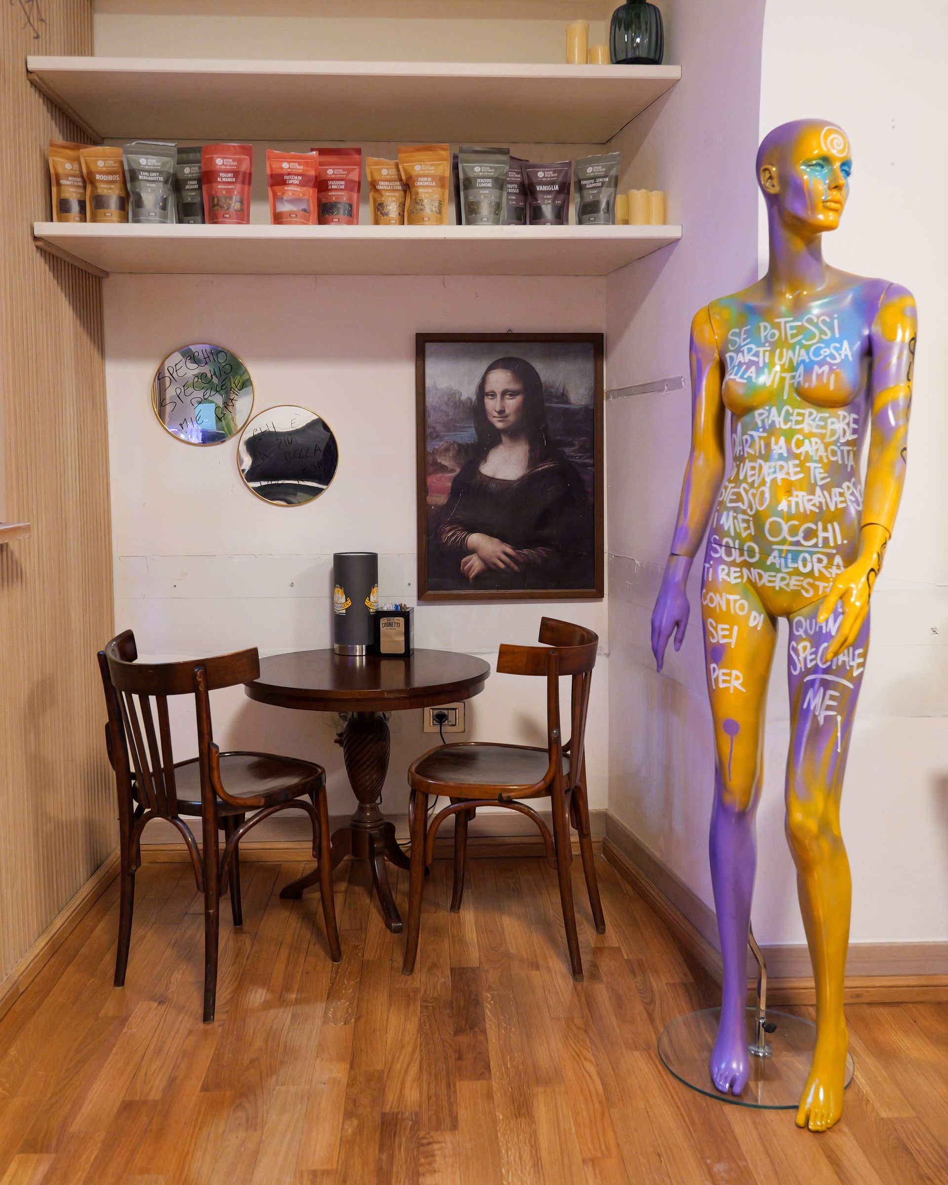 Interno con tavolino e sedie, un manichino e una stampa della Gioconda. Mensole con barattoli e specchi.