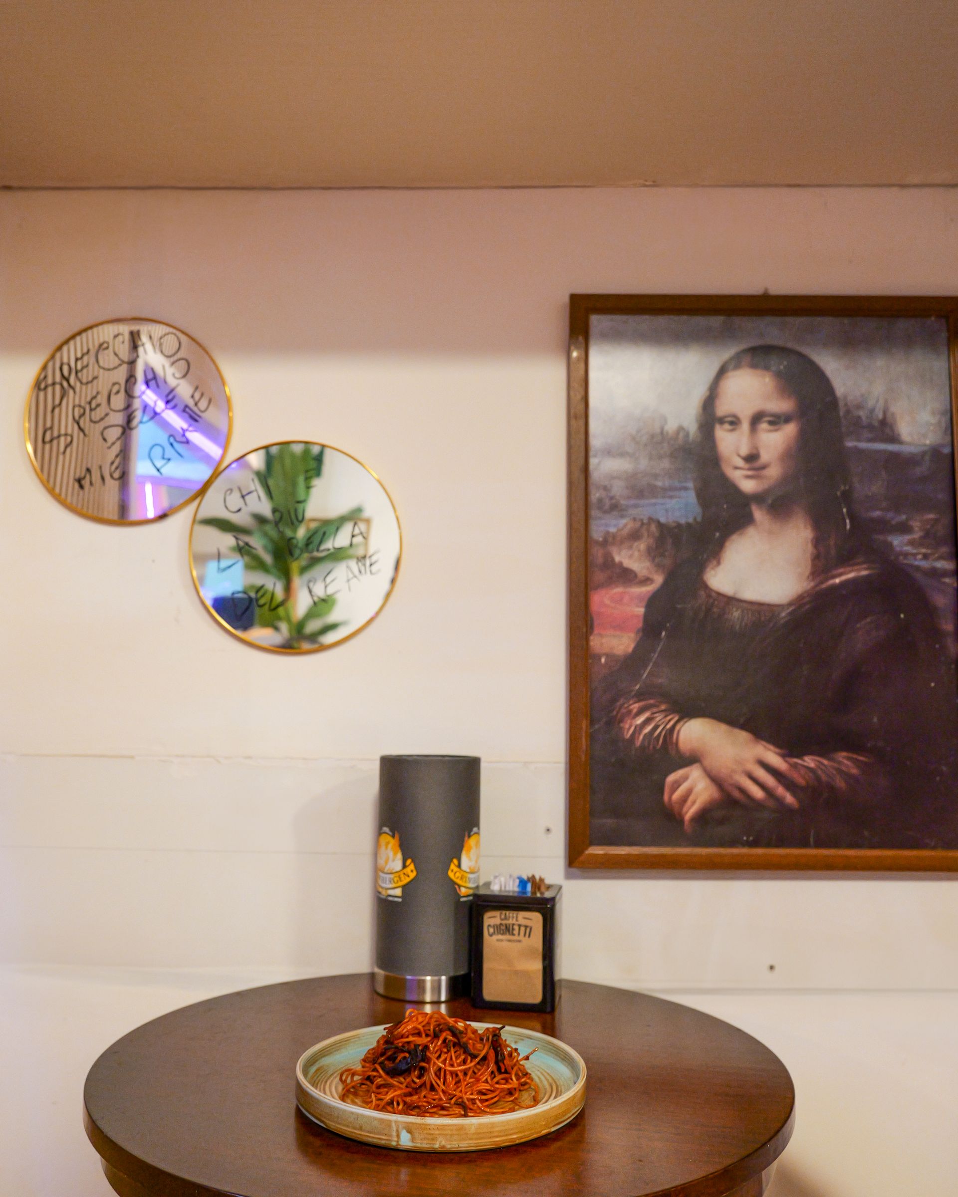 Tavolo rotondo con opere d'arte, una lampada e cibo sotto un dipinto della Gioconda.