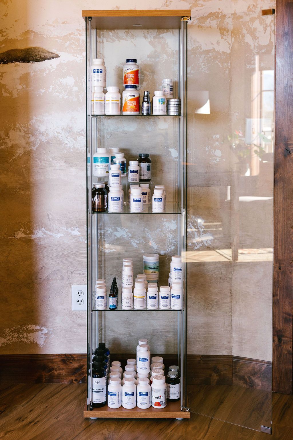 Glass cabinet filled with white supplement bottles against a textured wall and wooden floor.