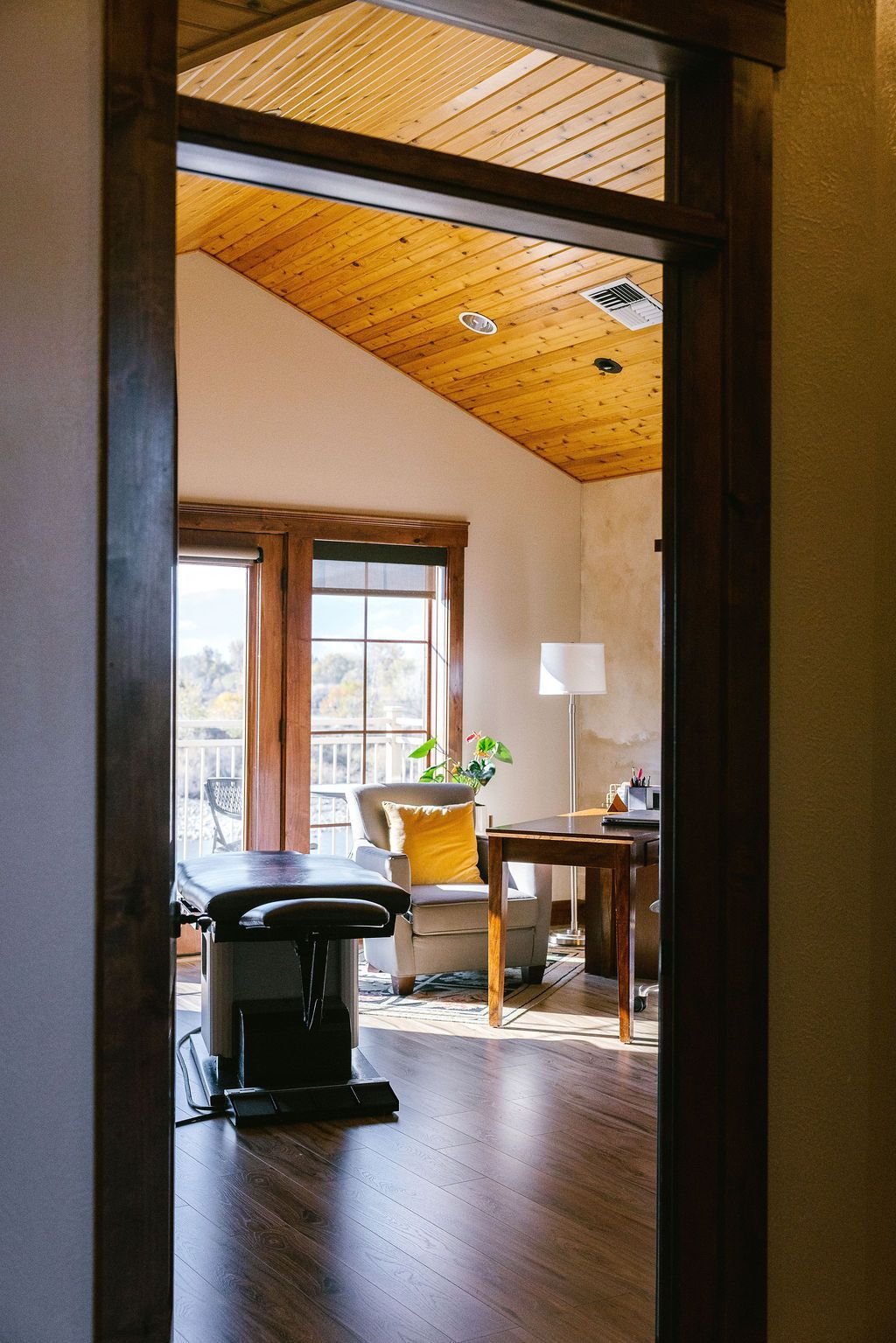 View through doorway into light-filled room with wood ceiling, window, desk, and chair.