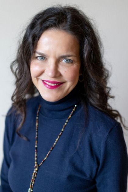A woman wearing a blue turtleneck and a necklace is smiling for the camera.