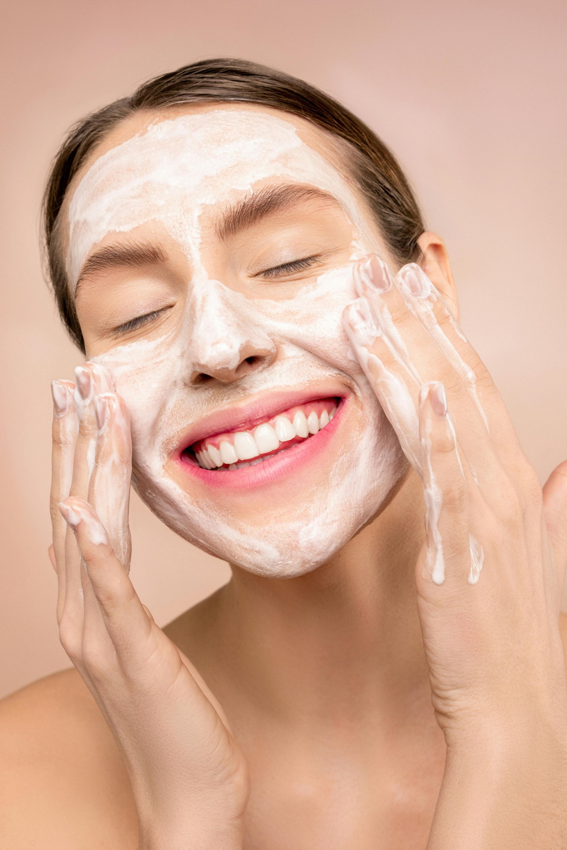 A woman is smiling while washing her face with a white mask.