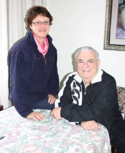 Two people smiling beside a bed, one seated and one standing, in a bright room.