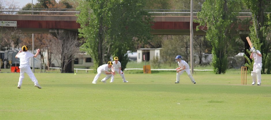 Cricket players fielding on a green pitch near a pavilion, with batsman at left and bowler at right