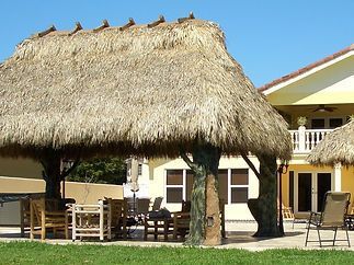 A thatched gazebo with chairs and tables in front of a house.
