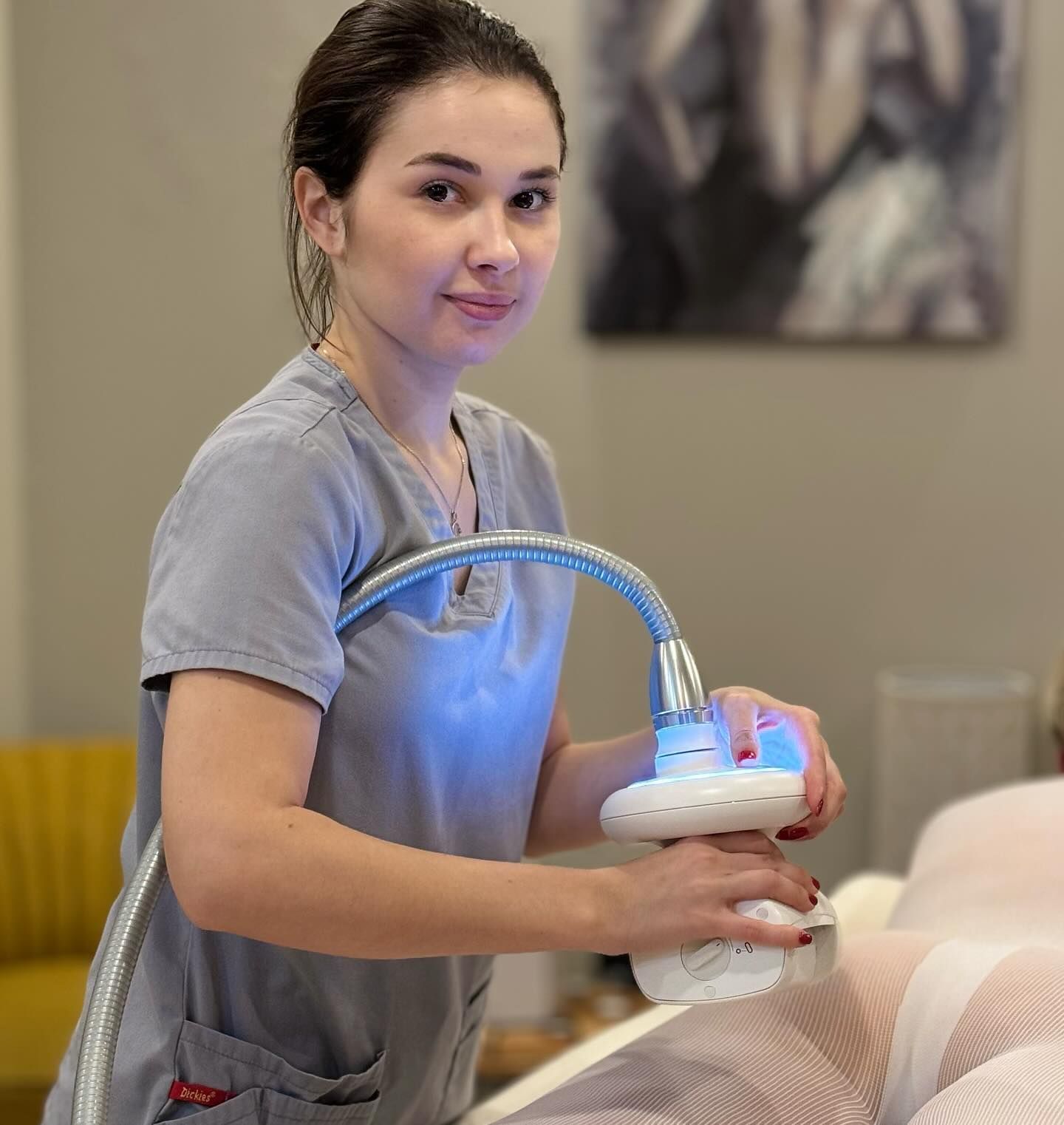 Woman in scrubs operates a medical device on a patient; setting is a treatment room.
