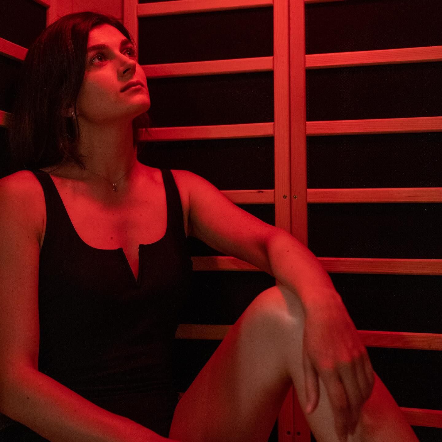 Woman in a sauna, looking upwards, lit by red light, wearing a black tank top.