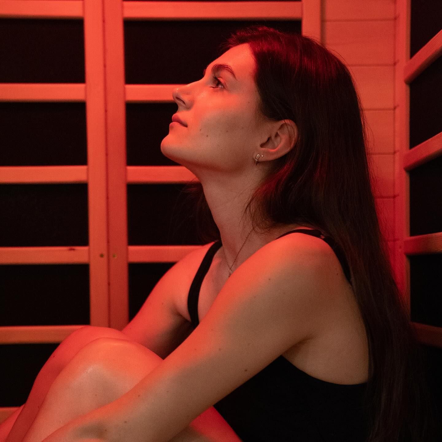 Woman in a red-lit sauna, looking upward with a contemplative expression.