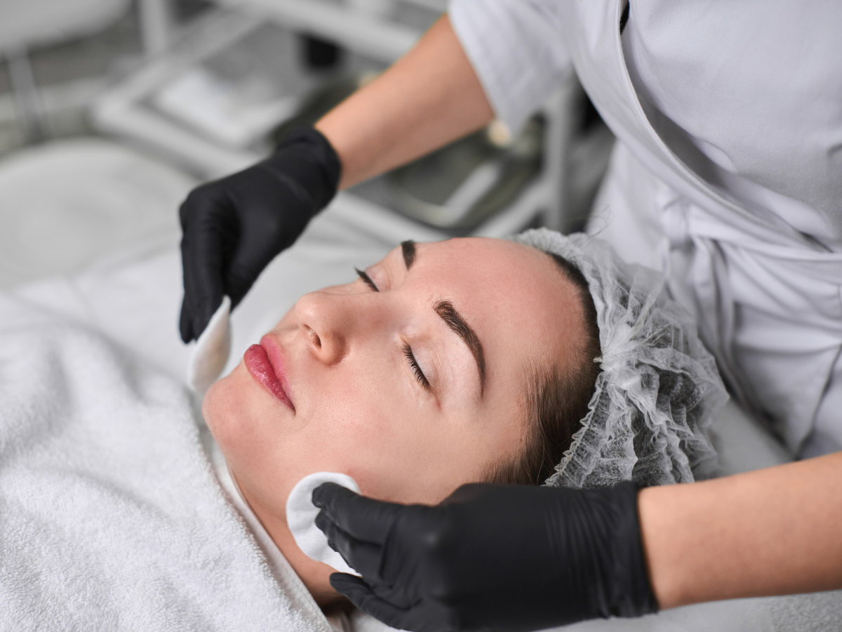 Woman receiving facial treatment; esthetician's hands in black gloves cleanse her face with cotton pads.
