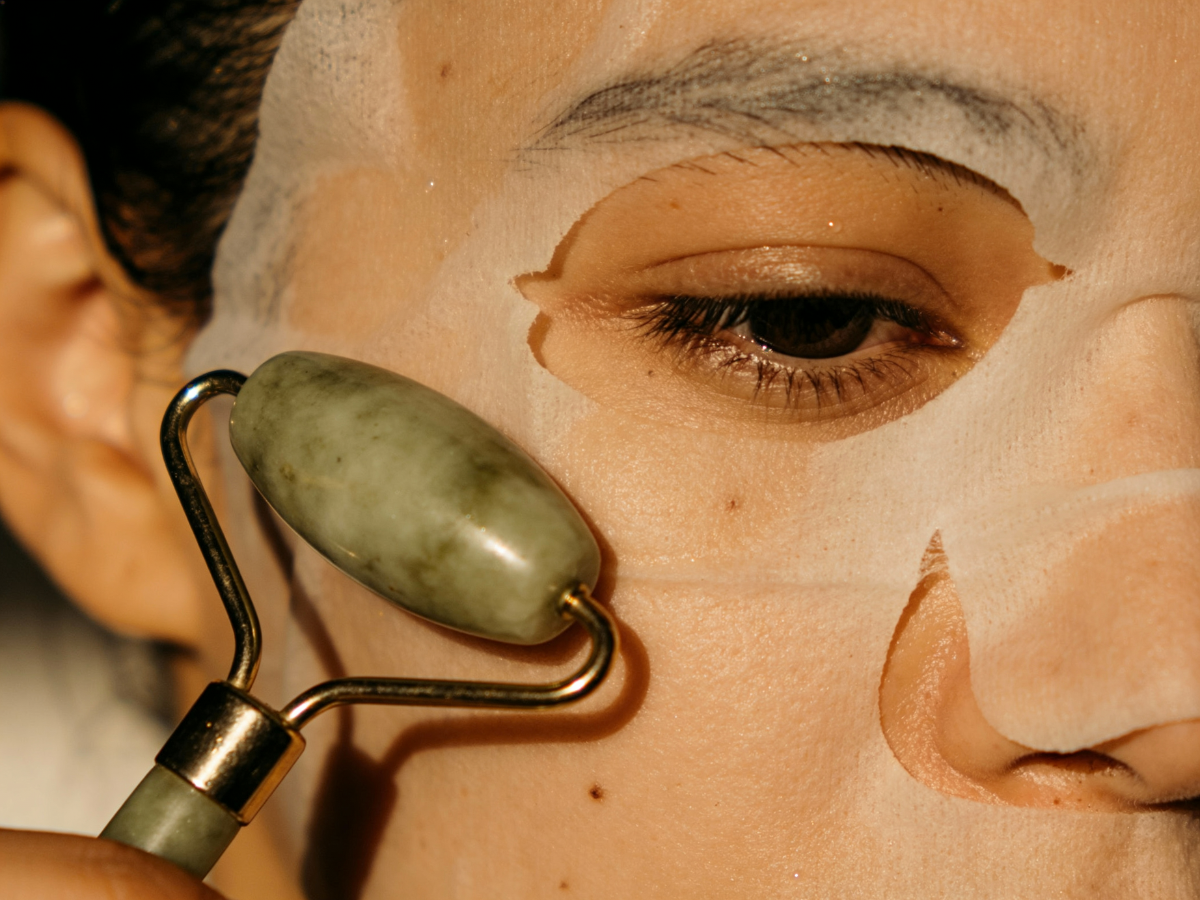 Person using a jade roller on their face while wearing a sheet mask.
