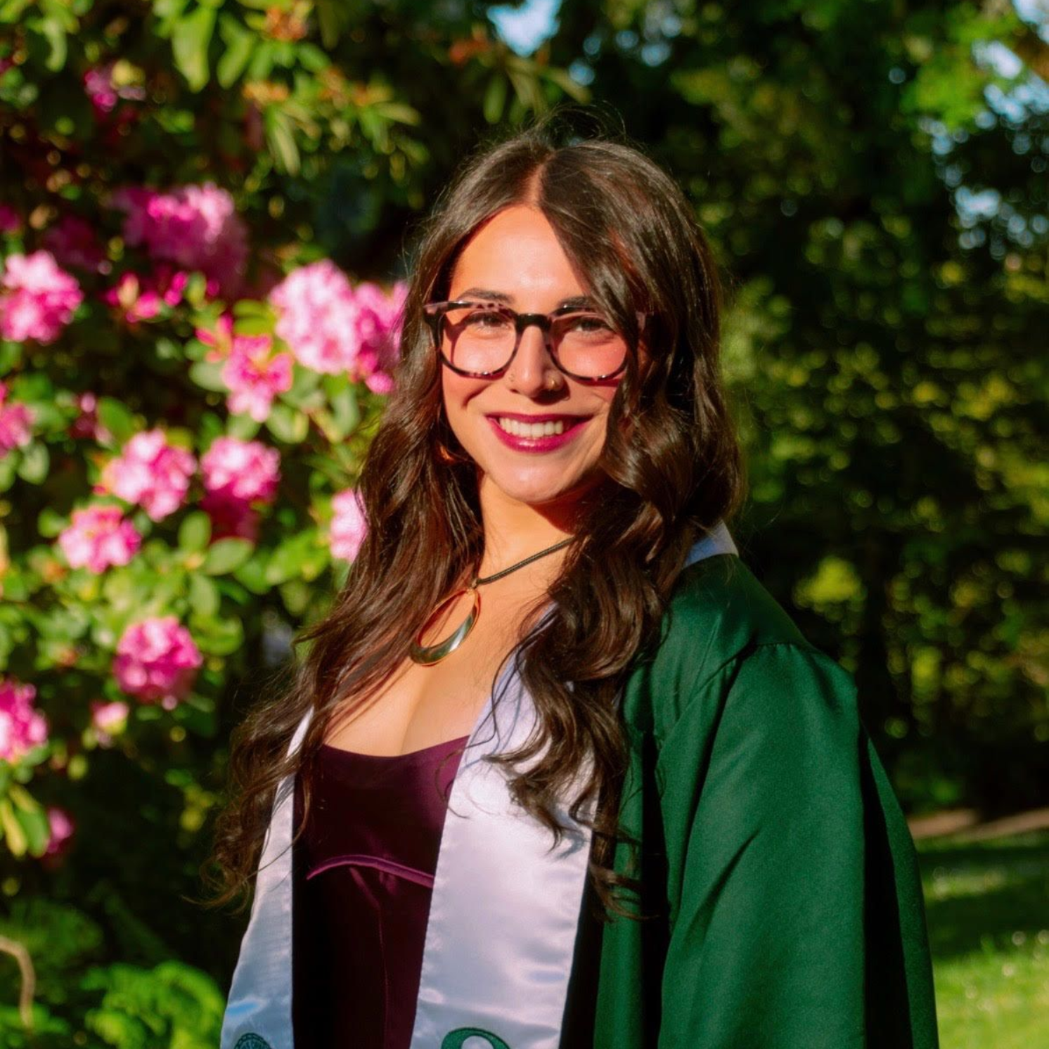 Woman in graduation gown smiles outdoors near pink flowers.