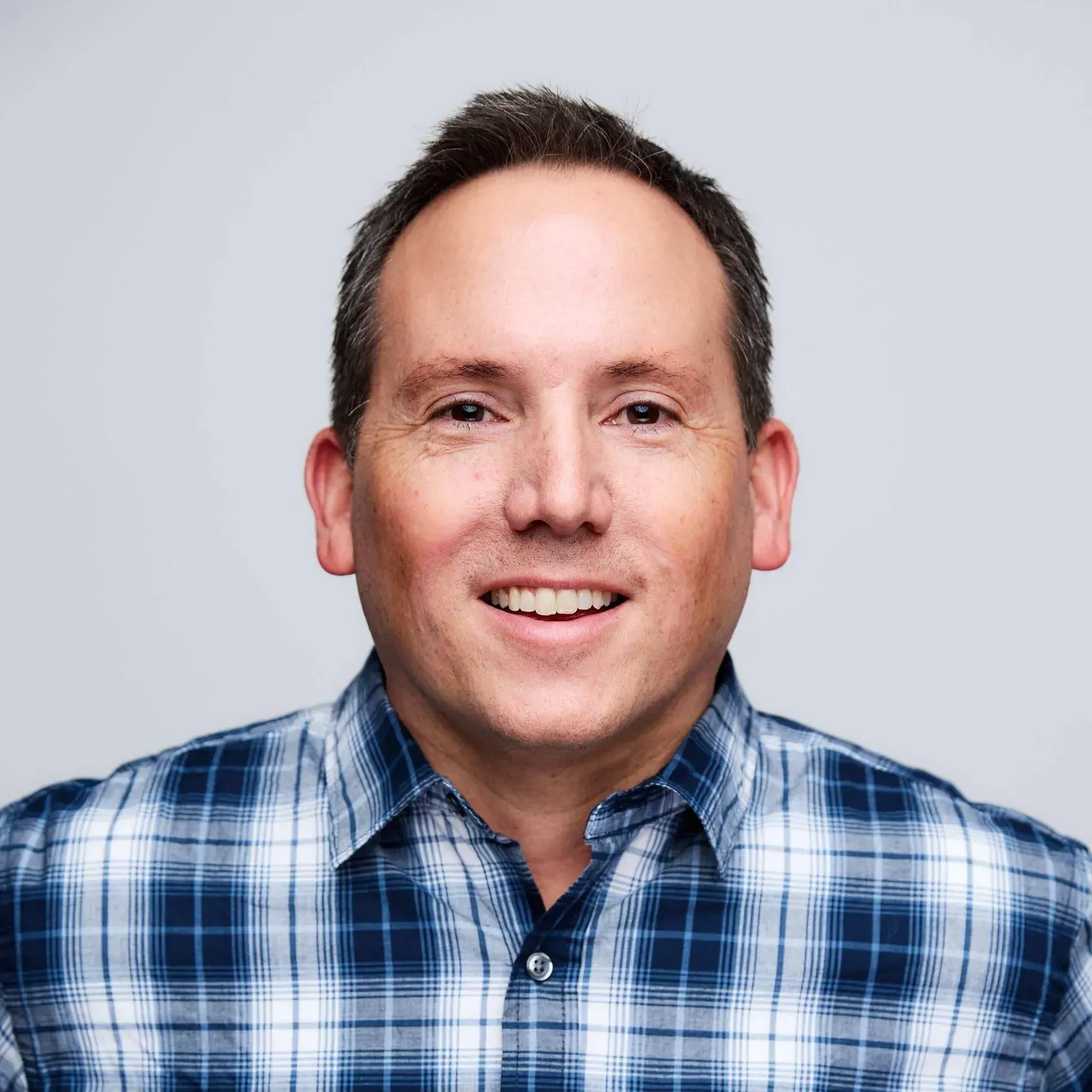 Man with short dark hair smiles, wearing a blue plaid shirt against a light gray background.