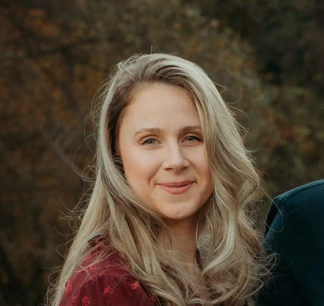 Blonde woman smiling, wearing a burgundy top. Outdoors with blurred trees in the background.