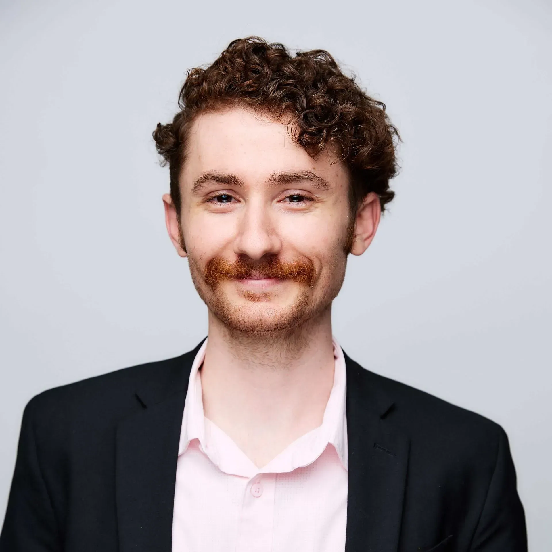Man with curly brown hair and a mustache wearing a pink shirt and black blazer.