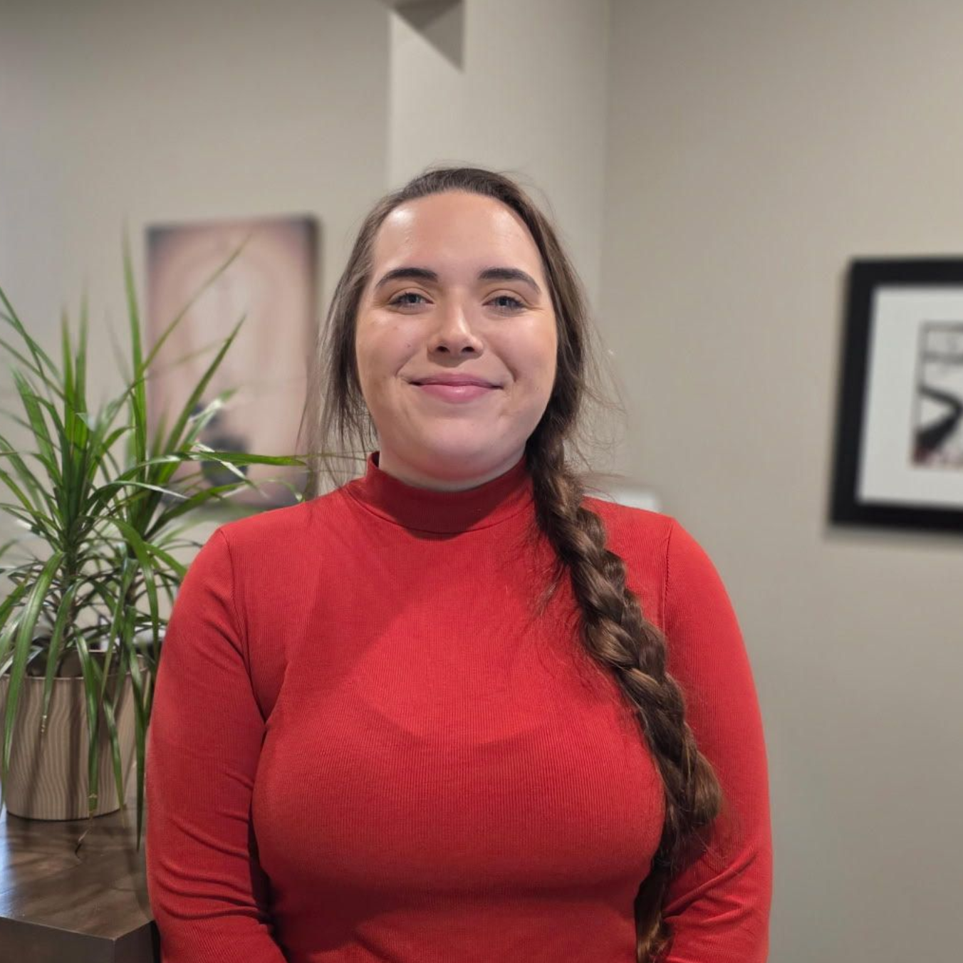 Woman with long braid, red turtleneck, smiles. Posed in room with plant, paintings.