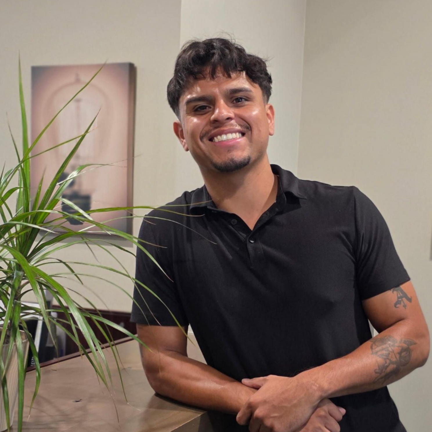 Man with a black shirt smiling leans on a counter next to a plant and artwork.