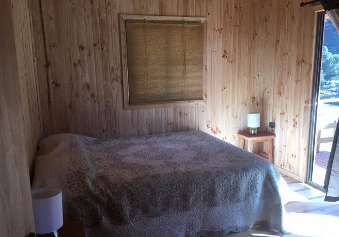 Dormitorio con paneles de madera, cama, mesita de noche, persiana y puerta abierta con vistas luminosas al exterior.