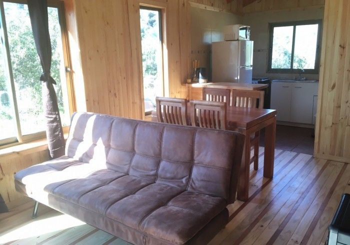 Interior de la cabaña revestido de madera con futón, mesa de comedor y zona de cocina junto a grandes ventanales.