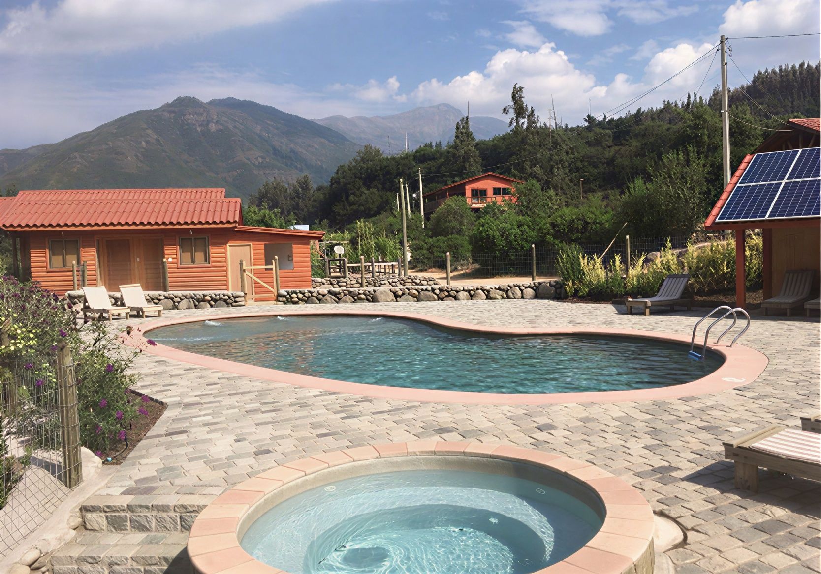 Piscina al aire libre y jacuzzi junto a una cabaña, con montañas y árboles al fondo.
