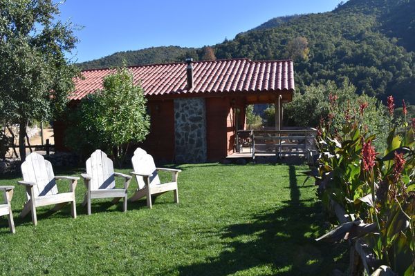 Sillas blancas sobre un césped verde junto a una cabaña de techo rojo con montañas al fondo.